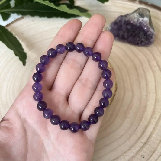 Bracelet Améthyste en perles violettes disposées sur une main, avec un géode d'améthyste en arrière-plan
