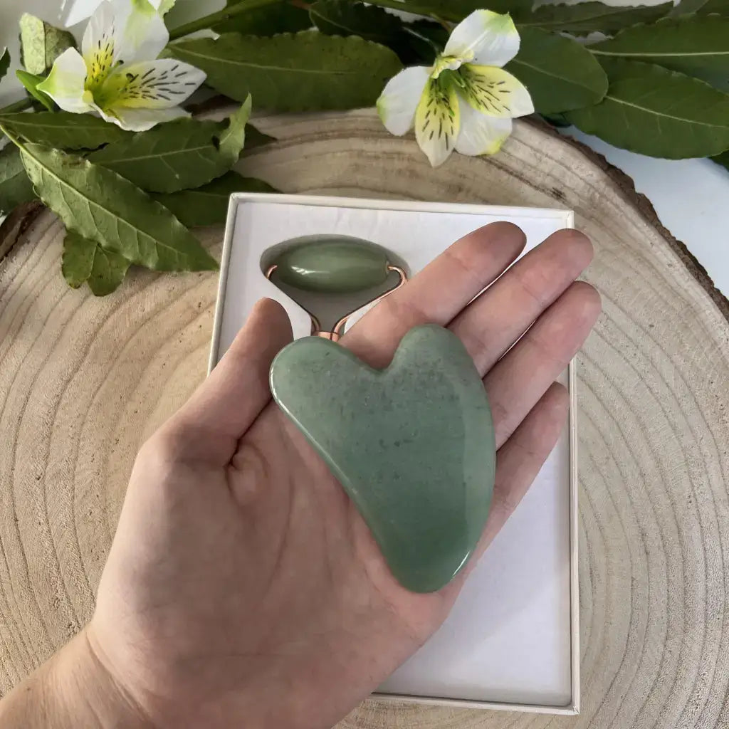 Gua sha et Rouleau de massage Aventurine verte dans une main, avec un fond naturel et des fleurs