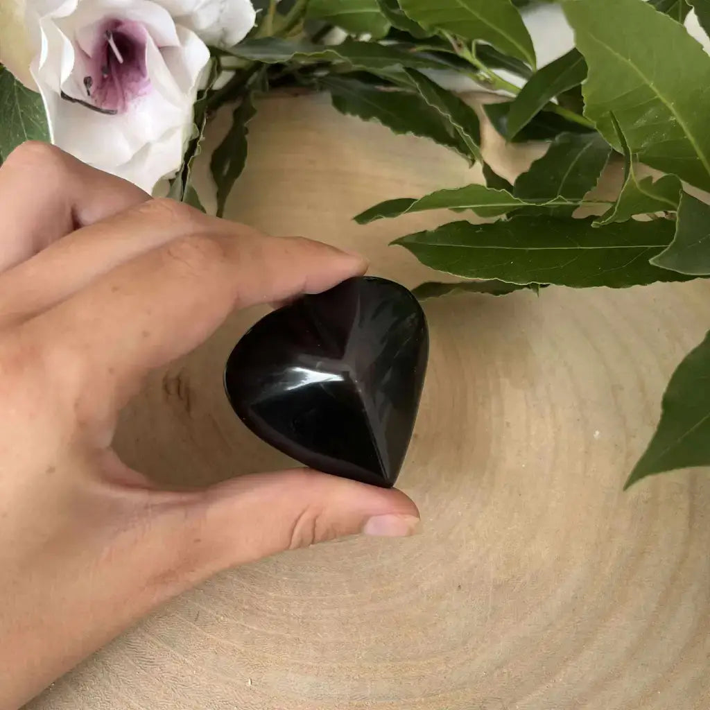 Galet en forme de cœur – Obsidienne Œil Céleste. Pierre polie noire tenue à la main sur un fond de bois avec feuillage décoratif