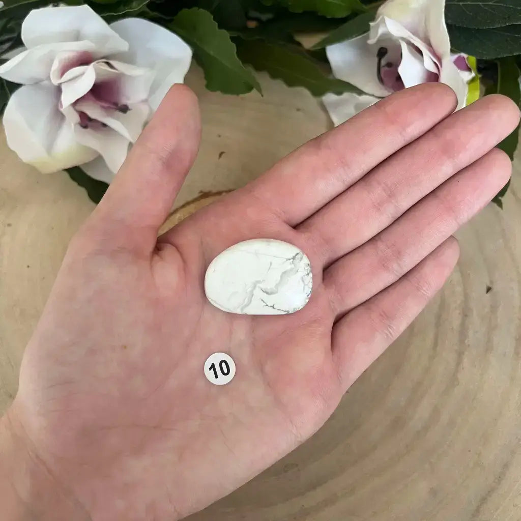 Galet Plat Howlite Blanche dans une main sur fond bois avec etiquette 10 et fleurs decoratives